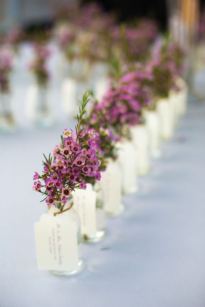 small vases filled with purple flowers are lined up on a long white tablecloth