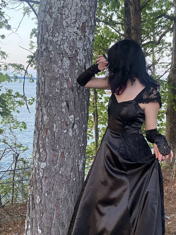 a woman in a black dress standing next to a tree and looking at the water