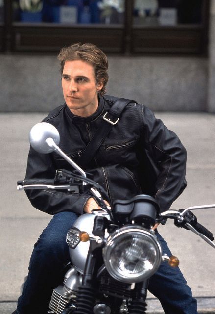 a man sitting on top of a motorcycle in front of a building with his hand on the handlebars