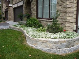 a stone retaining wall in front of a house with white flowers and greenery on the side