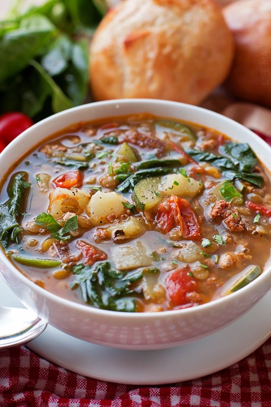 a bowl of soup with spinach, potatoes and other vegetables next to bread on a table