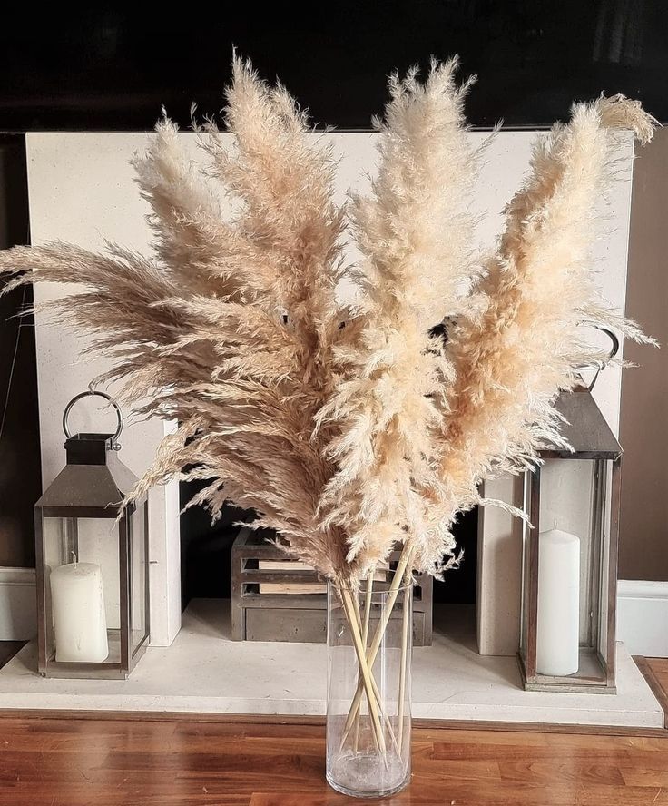 some dry grass in a glass vase on a wooden table next to lanterns and candles