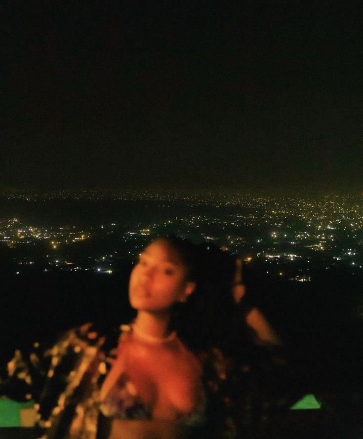 a woman standing on top of a hill at night with the city lights in the background