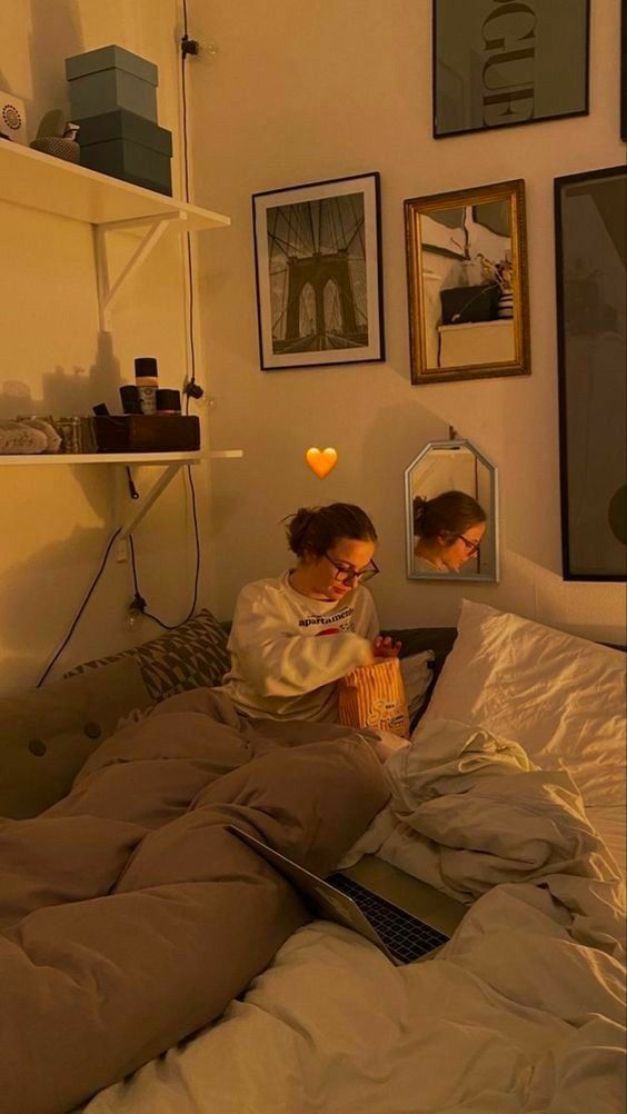 two women sitting on a bed looking at a laptop computer screen while one looks at her reflection in the mirror