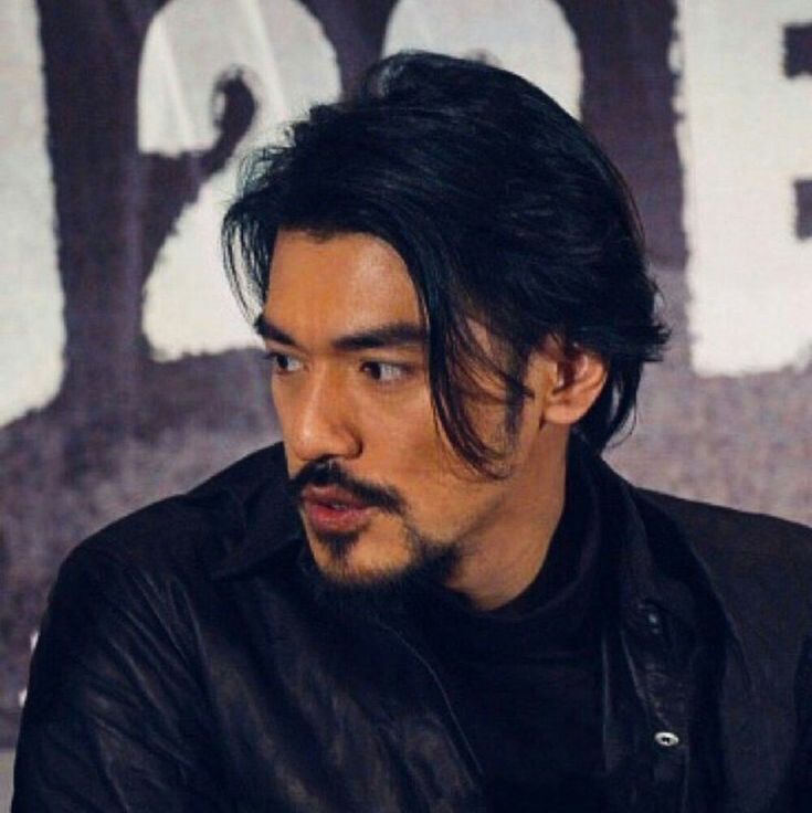 a man with black hair and a moustache looks at the camera while sitting in front of a poster