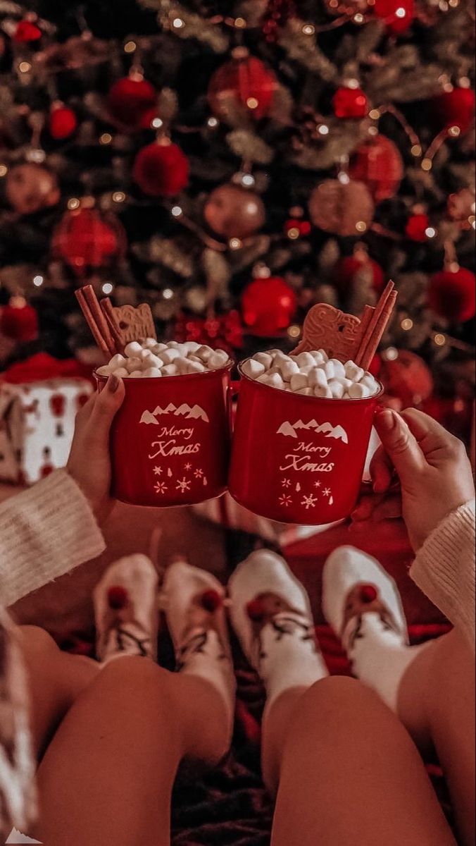 two people holding red mugs filled with marshmallows in front of a christmas tree