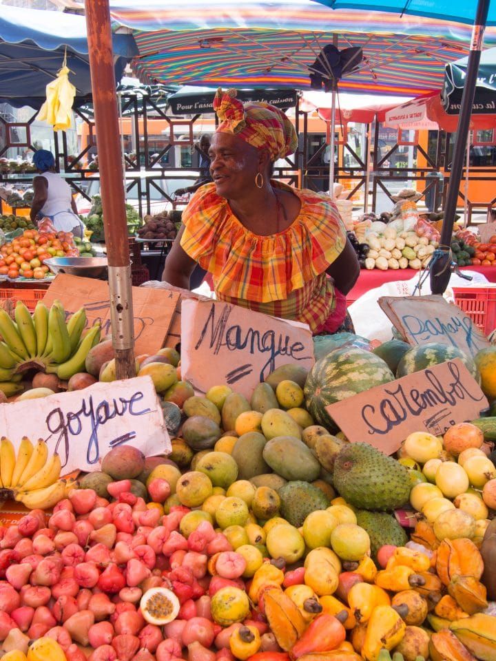 a woman standing in front of a pile of fruit at a farmers market with price signs