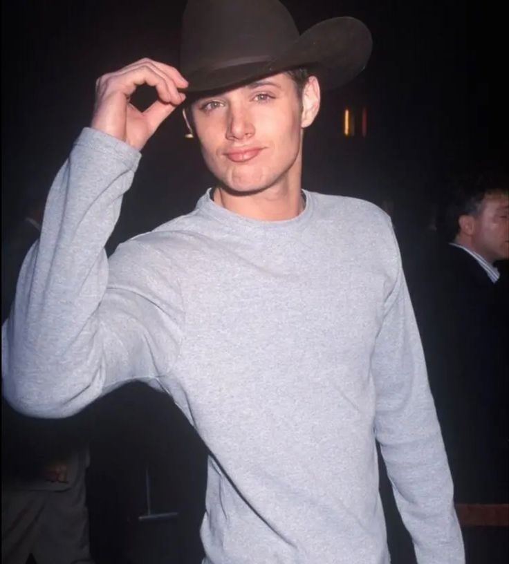 a young man wearing a cowboy hat and grey sweater posing for the camera with his hand on his head