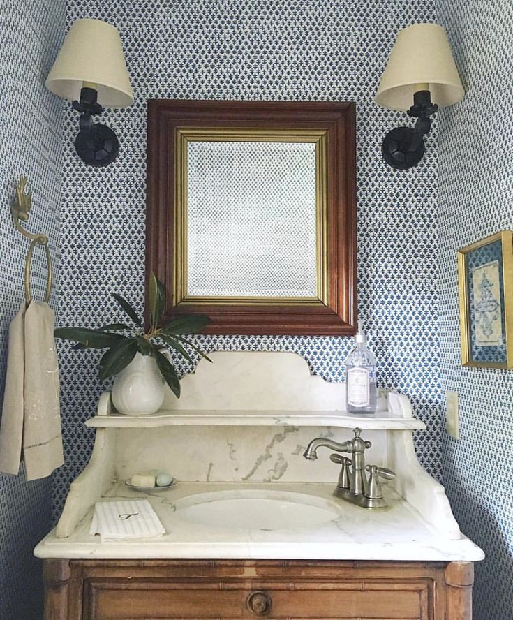 a bathroom with blue and white wallpaper has a wooden cabinet under the vanity mirror