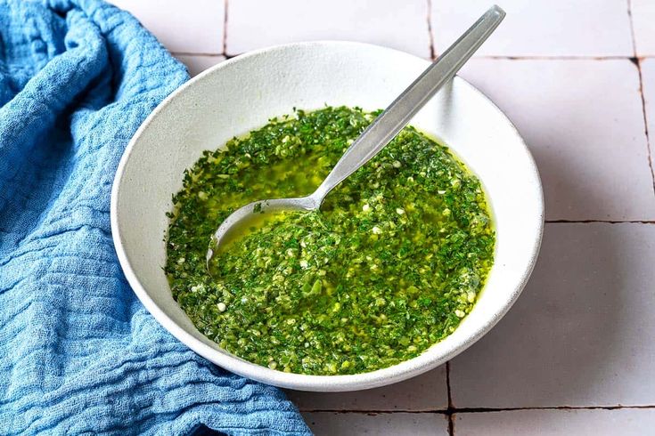 a white bowl filled with pesto on top of a blue towel next to a spoon