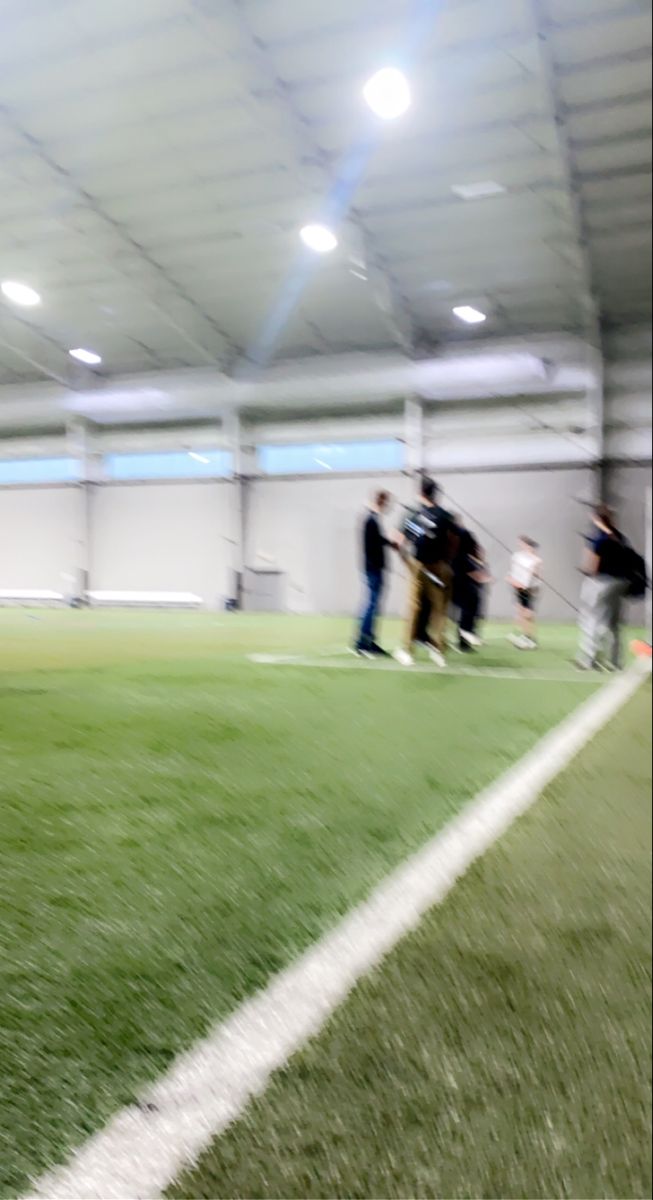 blurry photograph of people playing baseball in an indoor batting area with grass and white walls