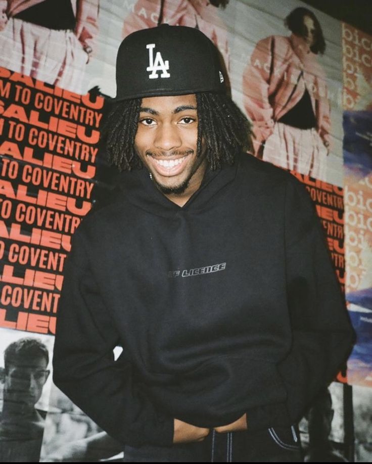 a man with dreadlocks and a baseball cap standing in front of a wall