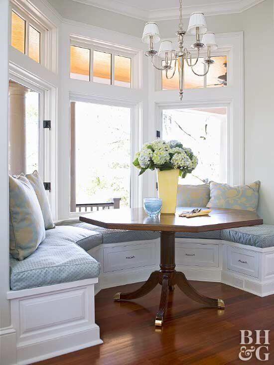 a table with some flowers on it in front of a bay window and bench that is built into the wall