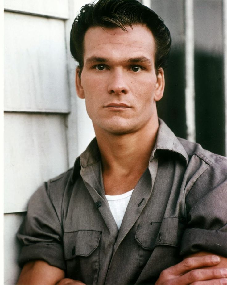 a man leaning against a wall with his arms crossed and looking at the camera while wearing a gray shirt