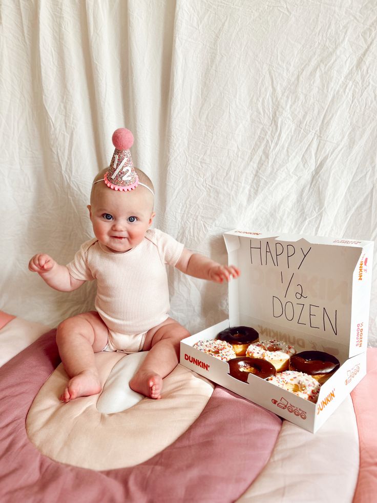 a baby in a birthday hat sitting next to a box of doughnuts that says happy v's dozen