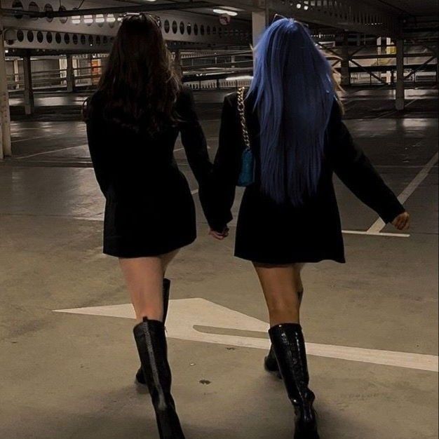 two women dressed in short black dresses and boots walking down a parking lot holding hands