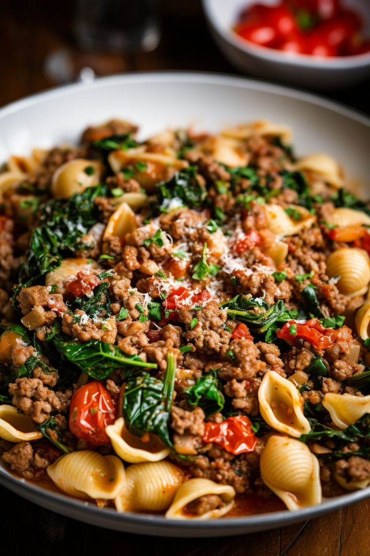 pasta with meat and spinach in a white bowl on a wooden table next to tomatoes