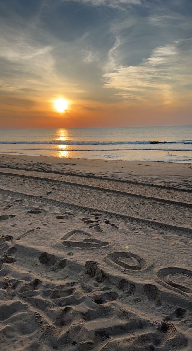 the sun is setting over the beach with tracks in the sand and footprints in the sand