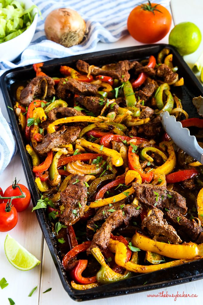 steak fajita with peppers and onions on a sheet pan next to tomatoes, limes, onion wedges