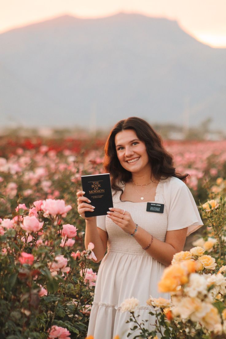 a woman standing in a field with flowers holding a book and smiling at the camera