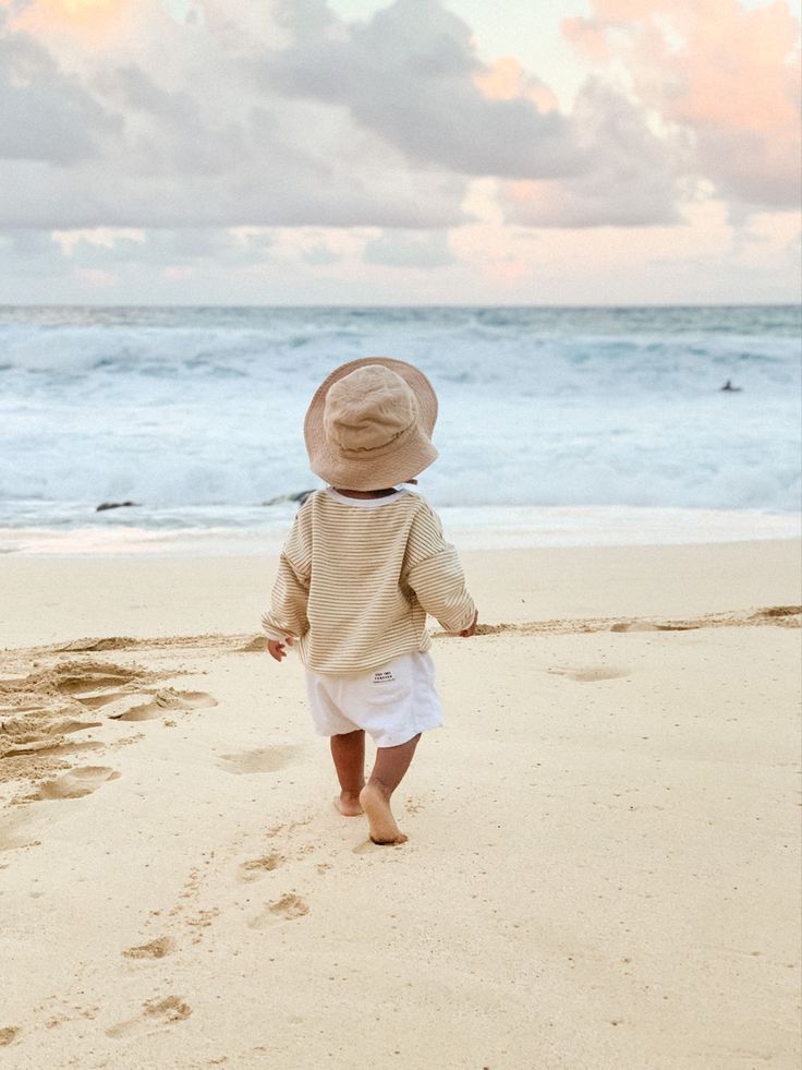 a little boy walking on the beach towards the ocean with his hat over his head