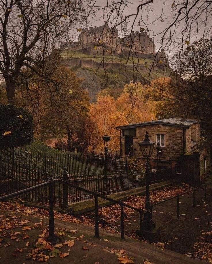 an old stone house in front of a castle with autumn leaves on the ground and trees around it