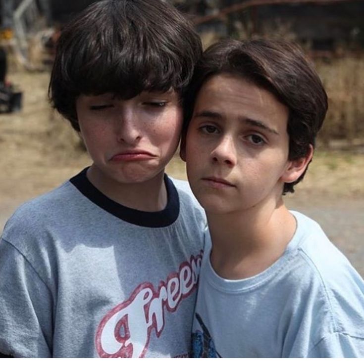 two young boys standing next to each other in front of a dirt field and building