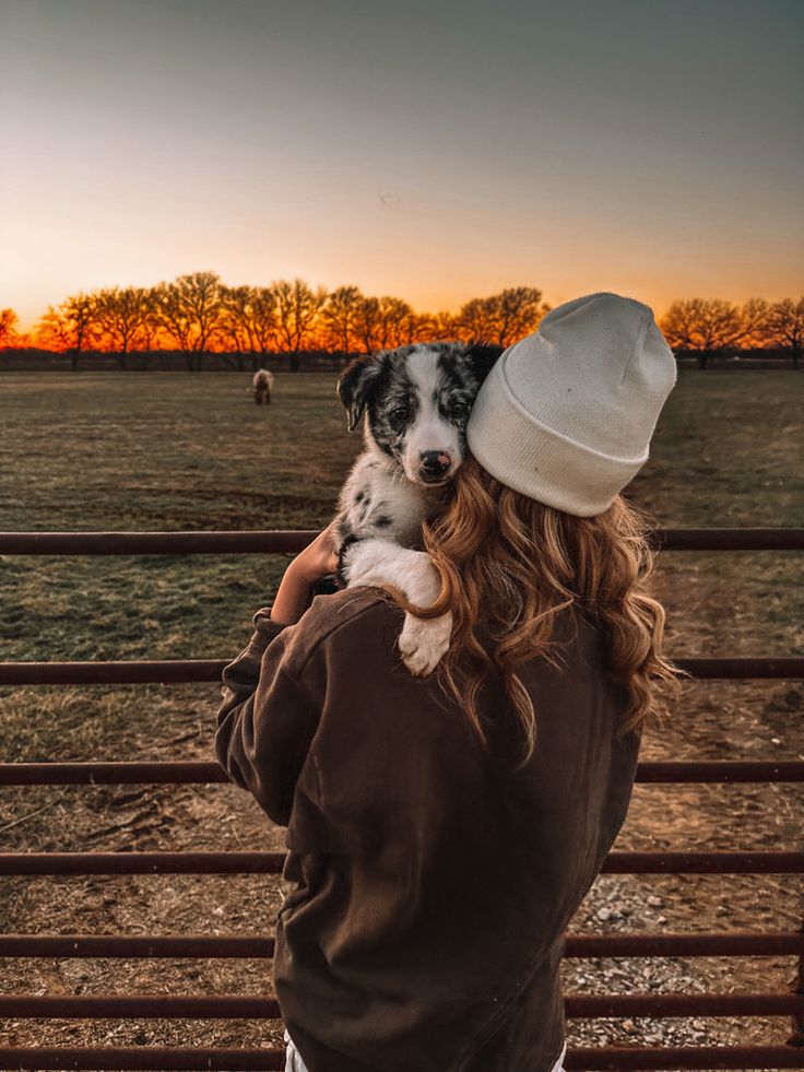 a woman holding a small dog in her arms while standing next to a fence at sunset