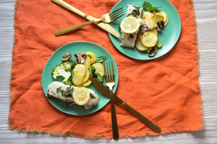 two plates with fish, lemons and broccoli are on an orange towel