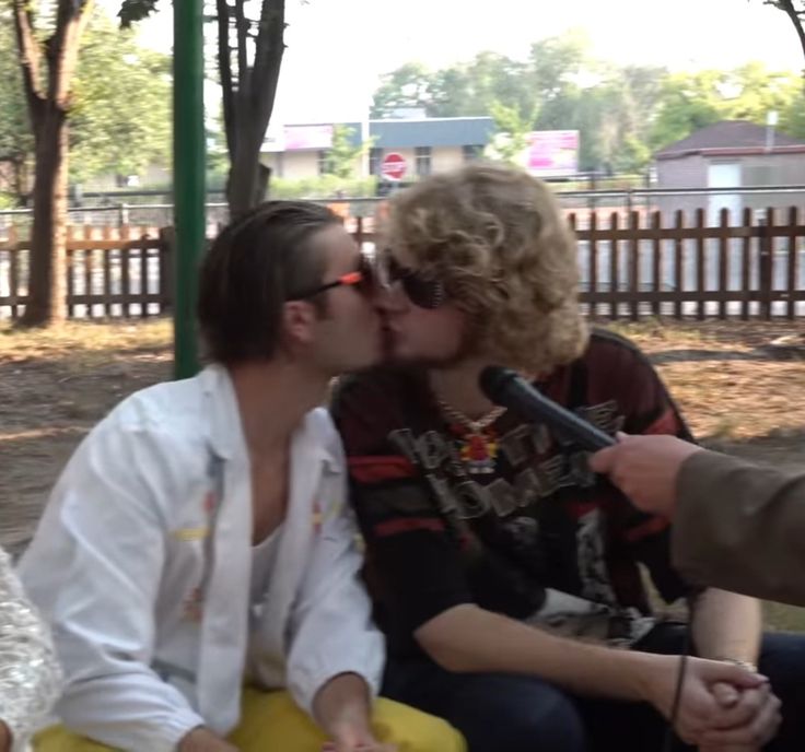 two people sitting on a bench and one person kissing the other with a microphone in front of them