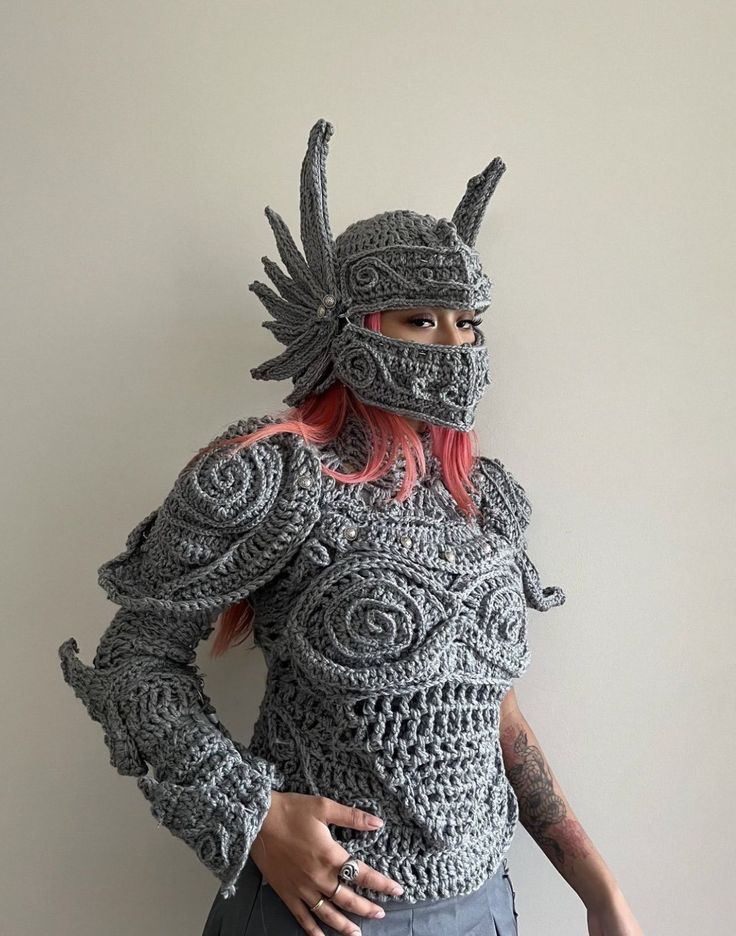 a woman with red hair wearing a crocheted costume and helmet, standing in front of a white wall