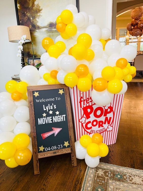 a popcorn box with yellow and white balloons in it next to a chalkboard sign