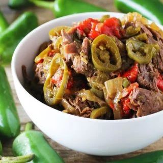 a white bowl filled with meat and peppers on top of a wooden table next to green peppers