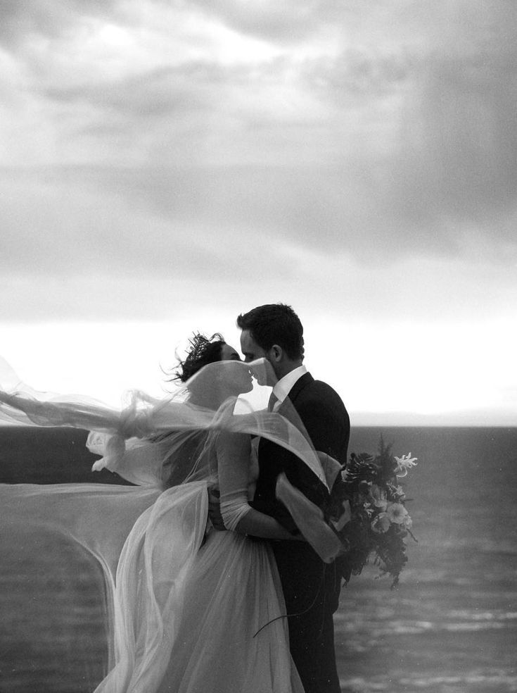 a bride and groom kissing in front of the ocean with their veil blowing in the wind