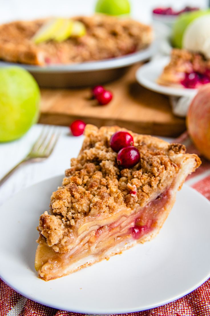 a slice of pie on a plate with apples and cranberries in the background