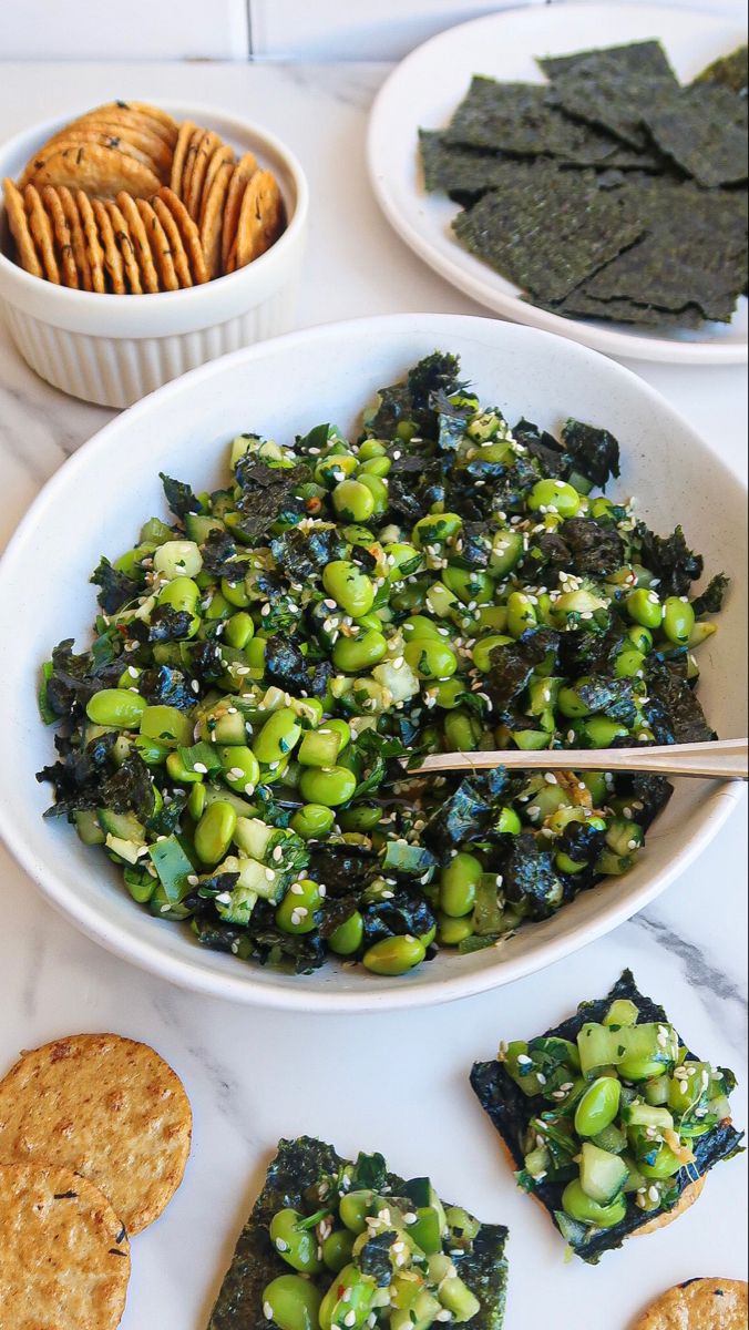 a bowl filled with green vegetables next to crackers and crackers on a table