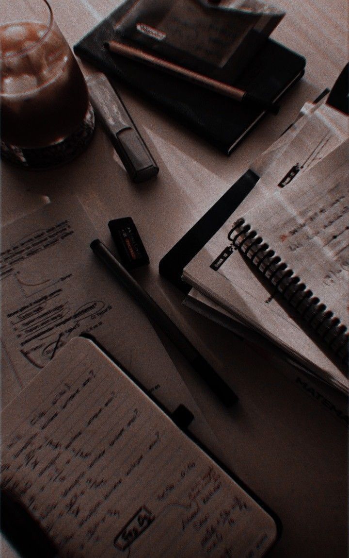 several notebooks and pens on a table with a glass of coffee in the background