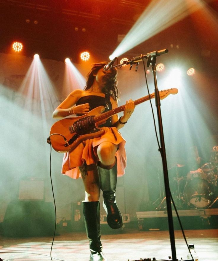 a woman with long hair and boots on stage holding a guitar in front of microphones