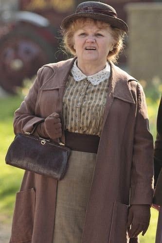 an older woman in a brown coat and hat walking down the street with her hand on her hip