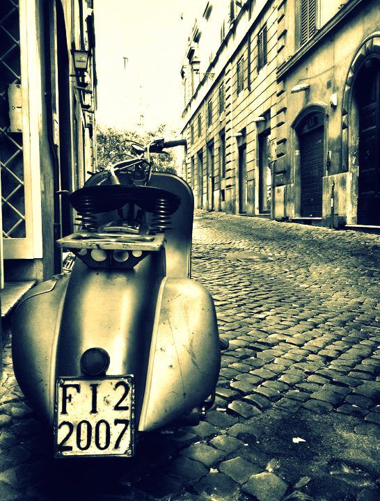 an old scooter parked on the side of a cobblestone road in front of a building