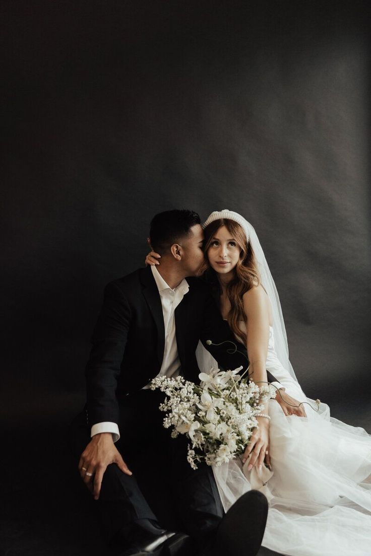 a bride and groom pose for a wedding photo in front of a dark background with their arms around each other