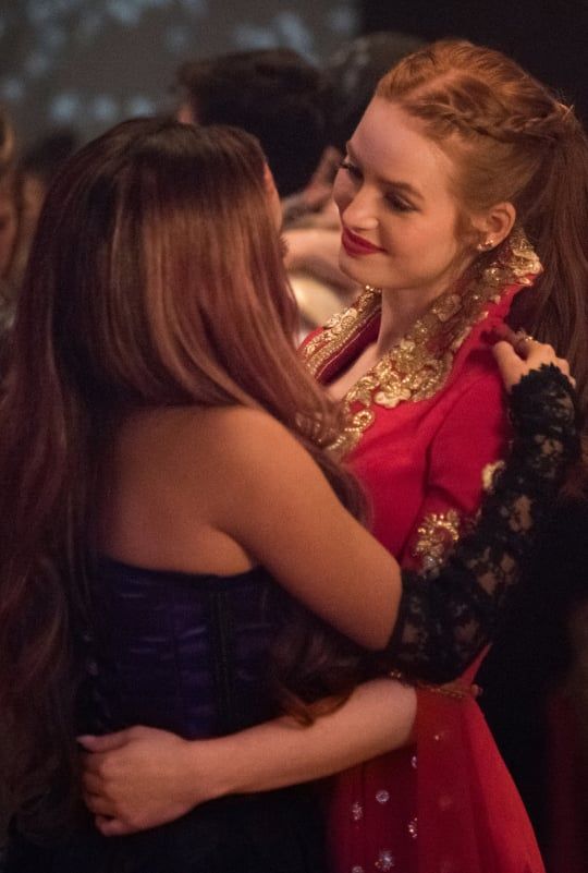 two young women embracing each other in a room full of people and one is wearing a red dress