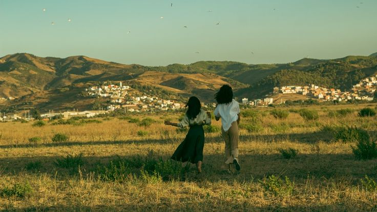 two women are walking in an open field with mountains in the background and birds flying overhead