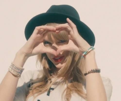 a woman wearing a hat and holding her hands up to her face while standing in front of a white wall