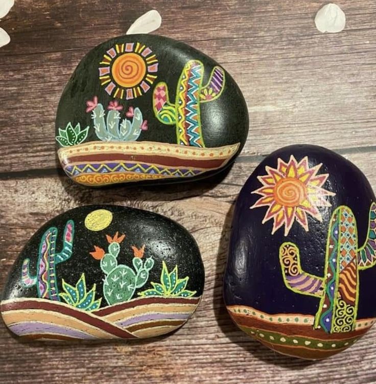 three painted rocks sitting on top of a wooden table next to flowers and cacti