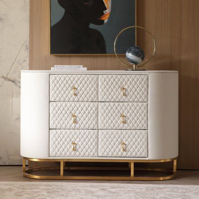 a white cabinet with gold handles and drawers in front of a painting on the wall