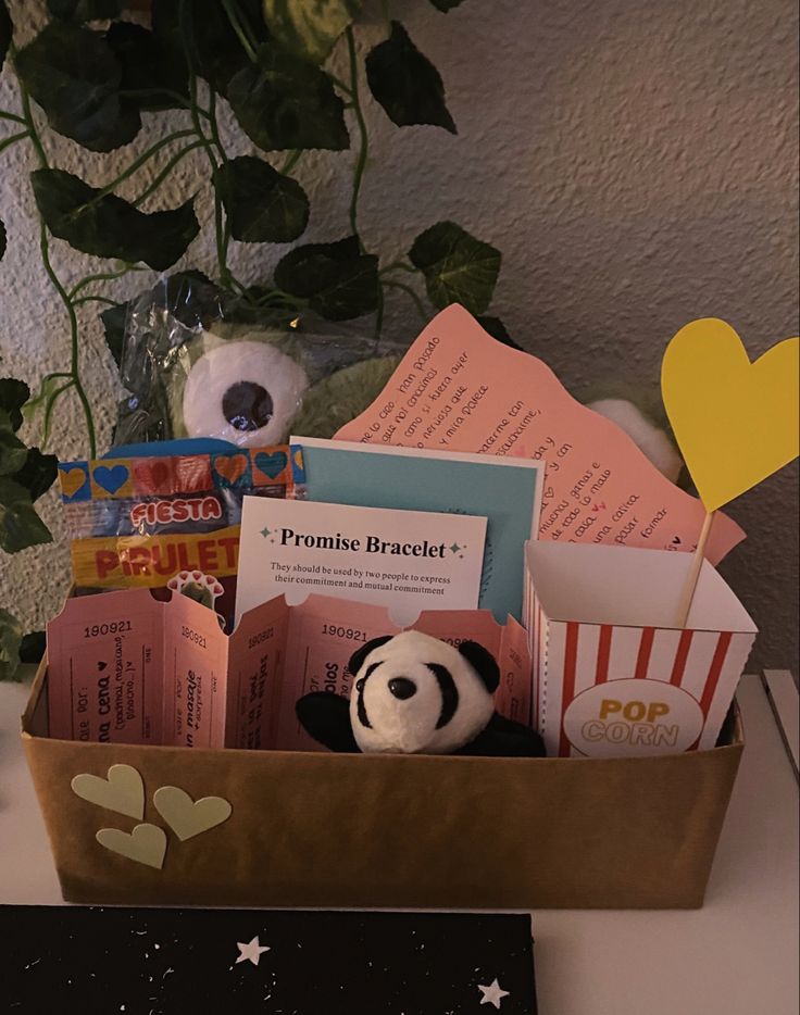 a stuffed panda bear sitting in a box filled with books and movie tickets next to a potted plant