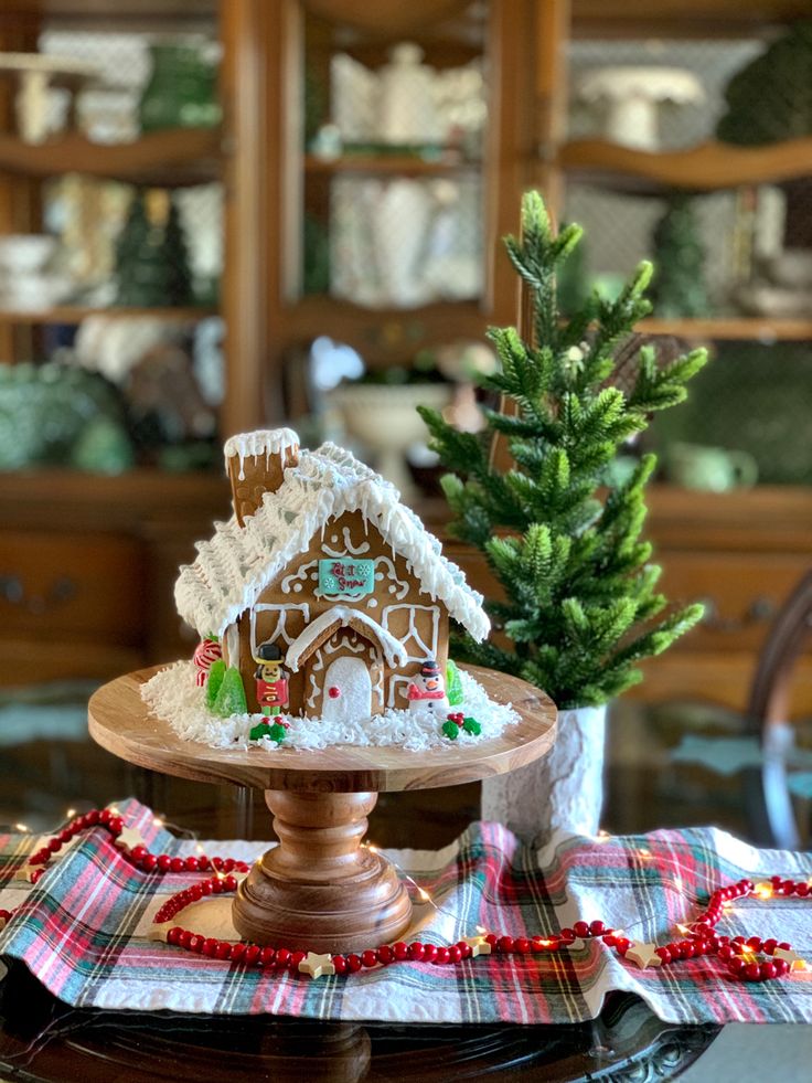 a gingerbread house on a cake plate next to a christmas tree