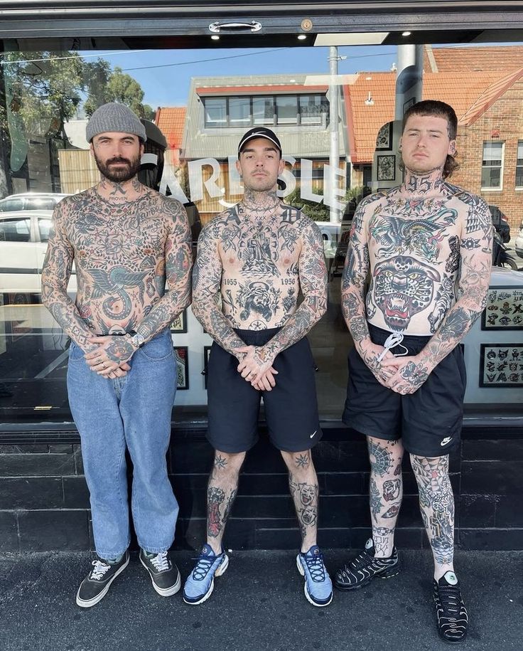 three men with tattoos standing in front of a store window, both looking at the camera