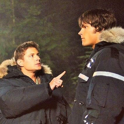 two men standing next to each other in front of trees and one pointing at something